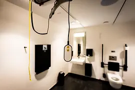 Interiorul unei toalete accesibile, cu sistem de ridicare montat în tavan și telecomandă suspendată, un lavoar cu oglindă și bare de sprijin lângă WC.