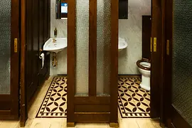 Vedere din interiorul unei toalete publice istorice, cu uși din lemn închis la culoare, ferestre din sticlă mată, pardoseală din plăci cu model și lavoare vintage în fața panourilor de marmură.