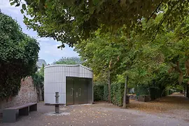 O clădire modernă de toaletă publică, cu fațadă metalică, de-a lungul unei alei mărginite de copaci în Augarten. Frunze uscate acoperă pe alocuri solul, iar bănci și garduri vii completează atmosfera liniștită a parcului.