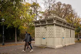 Un pavilion WC istoric, gri-deschis, cu elemente decorative din grilaj și detalii din sticlă colorată, înconjurat de copaci de toamnă, în timp ce trecătorii trec pe lângă el.