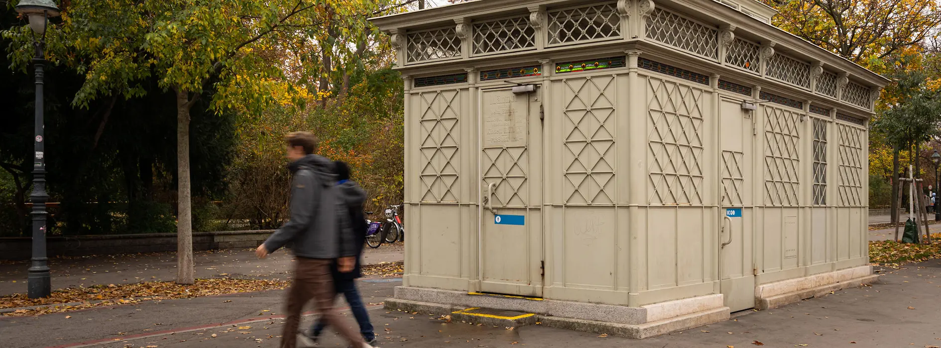 Un pavilion WC istoric, gri-deschis, cu elemente decorative din grilaj și detalii din sticlă colorată, înconjurat de copaci de toamnă, în timp ce trecătorii trec pe lângă el.