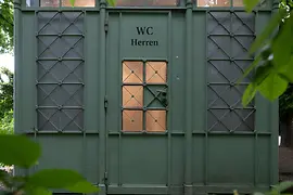 Un pavilion istoric de toaletă pentru bărbați, verde, cu grilaj metalic decorativ și ferestre din sticlă mată, parțial ascuns de frunziș luxuriant într-un peisaj de parc.