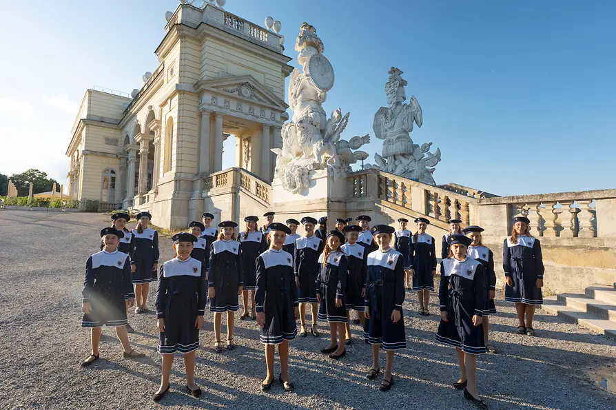 A bécsi kóruslányok egyenruhában állnak a Schönbrunni kastély parkjában lévő Gloriette előtt, történelmi épületekkel a háttérben.