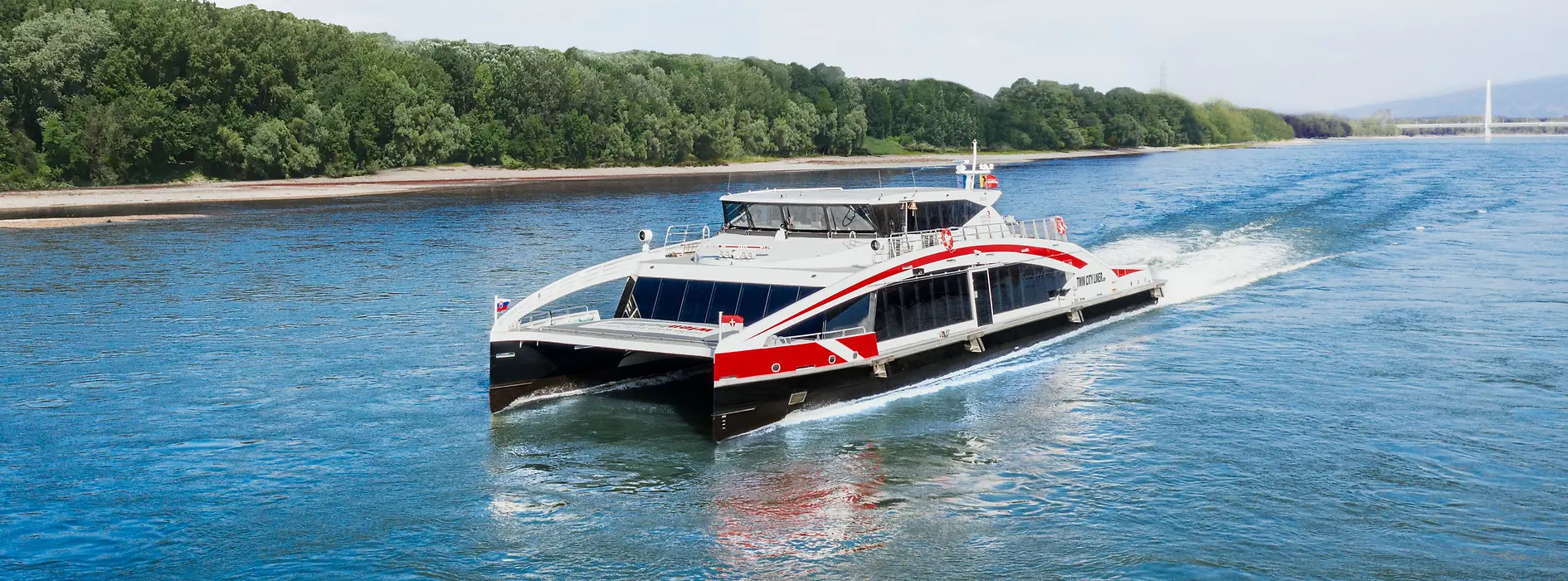 The high-speed catamaran Twin City Liner sails on the Danube with alluvial forest in the background.