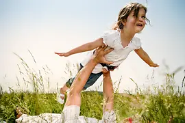 Adulto tumbado en la hierba que juega a que una niña pequeña vuela en el aire.