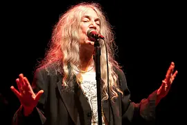 Patti Smith stands on a dark stage singing into a microphone.