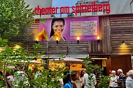 Exterior view of "Theater am Spittelberg" with red signage. A large poster featuring a smiling woman and event details is displayed on the wooden facade. People are gathered under umbrellas near the entrance, chatting.