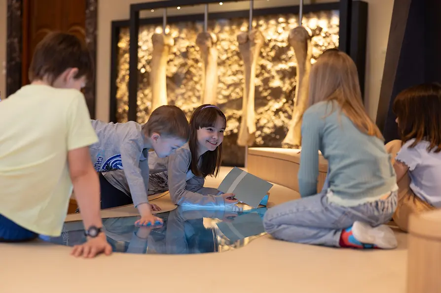 Kindereiszeitsaal: Fünf Kinder unterhalten sich an einer Touchpad-Station, im Hintergrund hängen riesige Knochen.