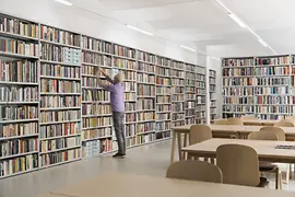 Library of the queer center Qwien with full bookshelves, tables, chairs, and a librarian placing a book back on the shelf