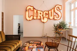 Reception area in the queer center Qwien with a sofa, a table and chairs, and a large neon sign for the Circus party series on the wall